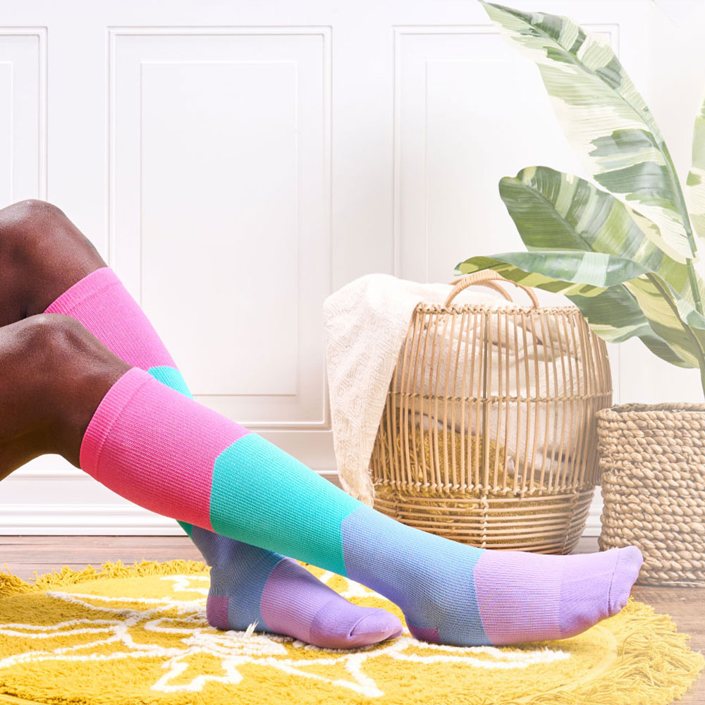 Woman wearing Color Block Bright Compression socks in multicolor, with color blocks of pink, turquoise, blue and purple, sitting out of frame with her feet on a bright yellow rug. A plant and a basket on in the background. 