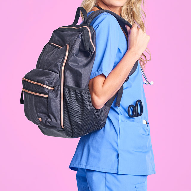 Woman wearing the Ultimate Nursing Backpack in grey on her back, with one hand on the strap. Woman is wearing light blue scrubs and has scissors in her pocket, all on a pink background