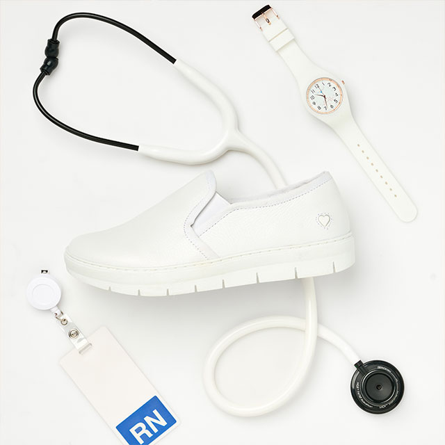 The Adela slip on sneaker in white on a white background, with a white and black stethoscope behind the shoe. There’s also a white watch in the upper right corner, and a white and blue “RN” tag in the lower left corner.