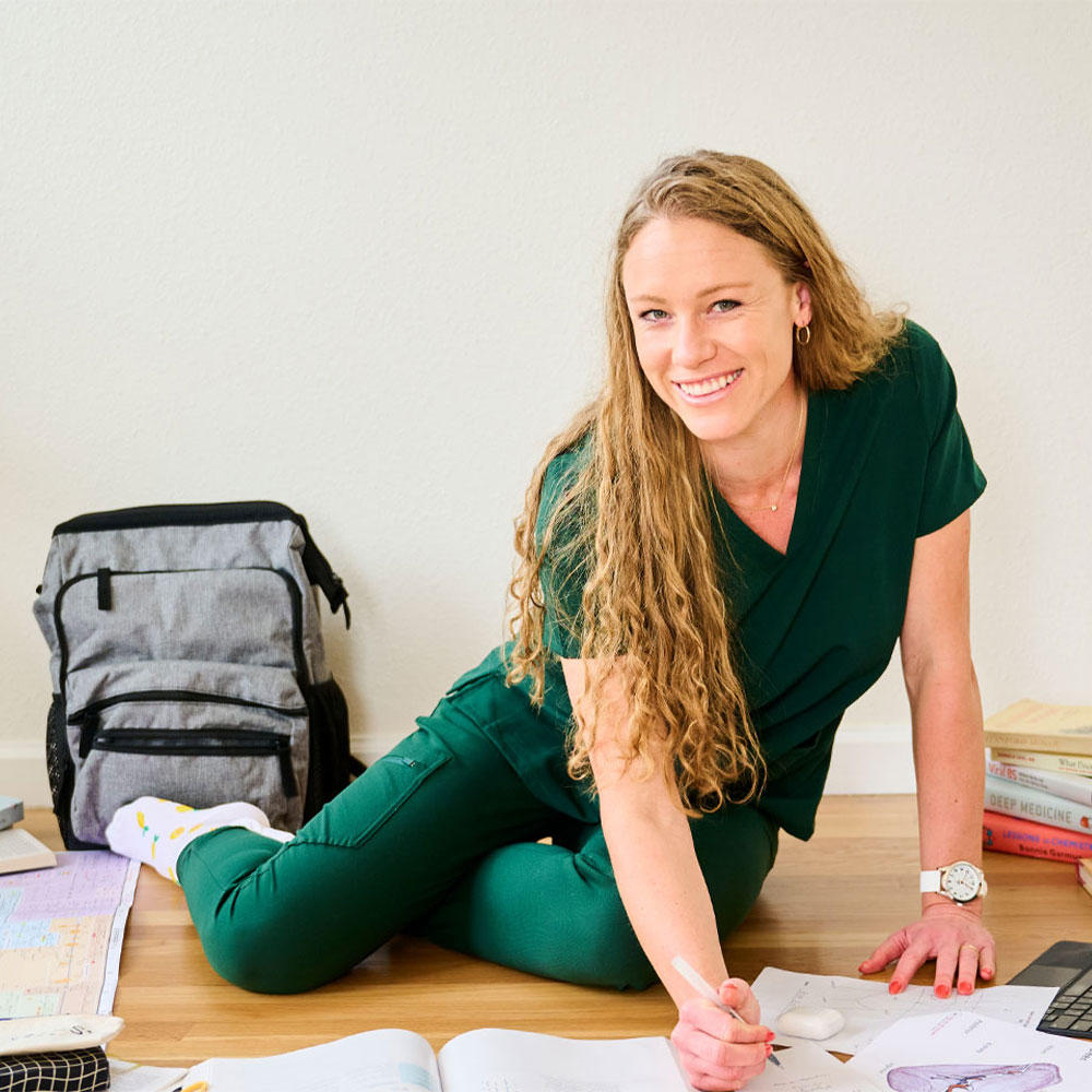 Nurse Mates Brand Partner Ella Eastin sitting on the floor surrounded by textbooks and study guides, with the Ultimate Nursing Backpack in grey behind her, and the Sweep Watch on her wrist. She’s wearing green scrubs and smiling at the camera.