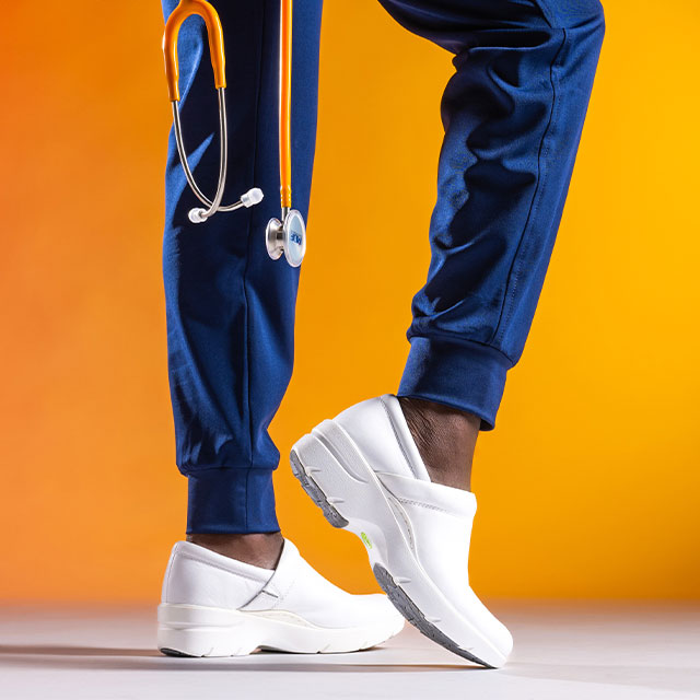 Woman wearing Align™ Indya clogs in white and dark blue scrub joggers  on an orange/yellow gradient background. An orange stethoscope falls into frame.