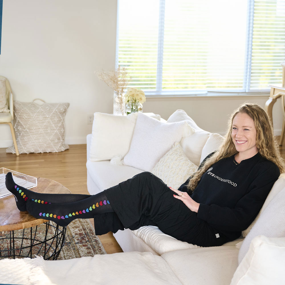 Nurse Mates Brand Partner Ella Eastin sitting on a couch wearing the Rainbow Hearts Compression Socks in black. 