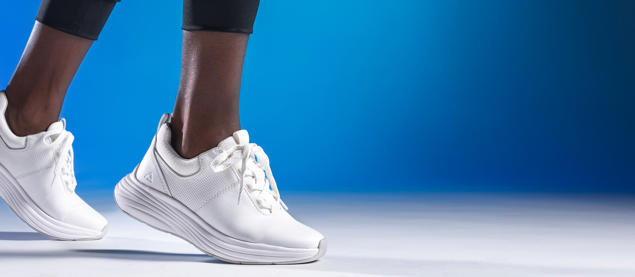 Woman wearing the Align™ Tulia in White, with her heels lifted slightly in a forward motion. Shot on a blue background