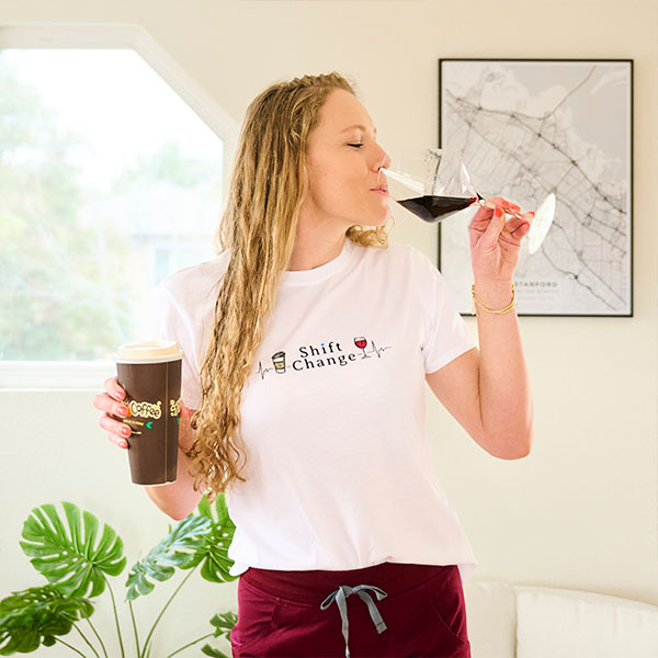 Ella Eastin, Nurse Mates brand partner, wearing the Shift Change Women’s Tee Shirt in white, hold a coffee in one hand, and motioning to take a sip of wine in the other hand. Shop Shift change shirt in white.