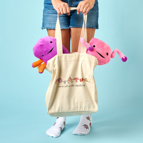 Woman holding the Organ Party canvas tote bag in tan in front of her, with two stuffed organs peeking out. Shop canvas tote bag in tan. 