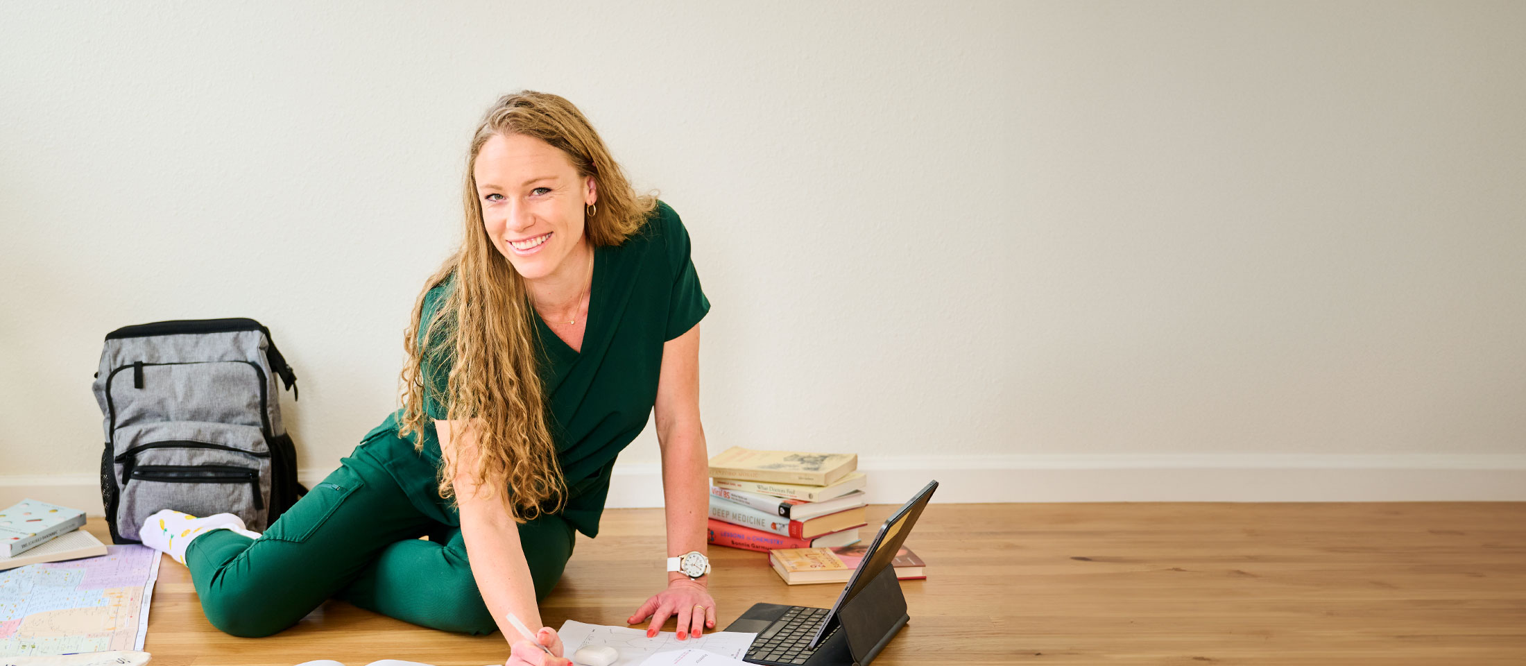 Nurse Mates Brand Partner Ella Eastin sitting on the floor surrounded by textbooks and study guides, with the Ultimate Nursing Backpack in grey behind her, and the Sweep Watch on her wrist. She’s wearing green scrubs and smiling at the camera.