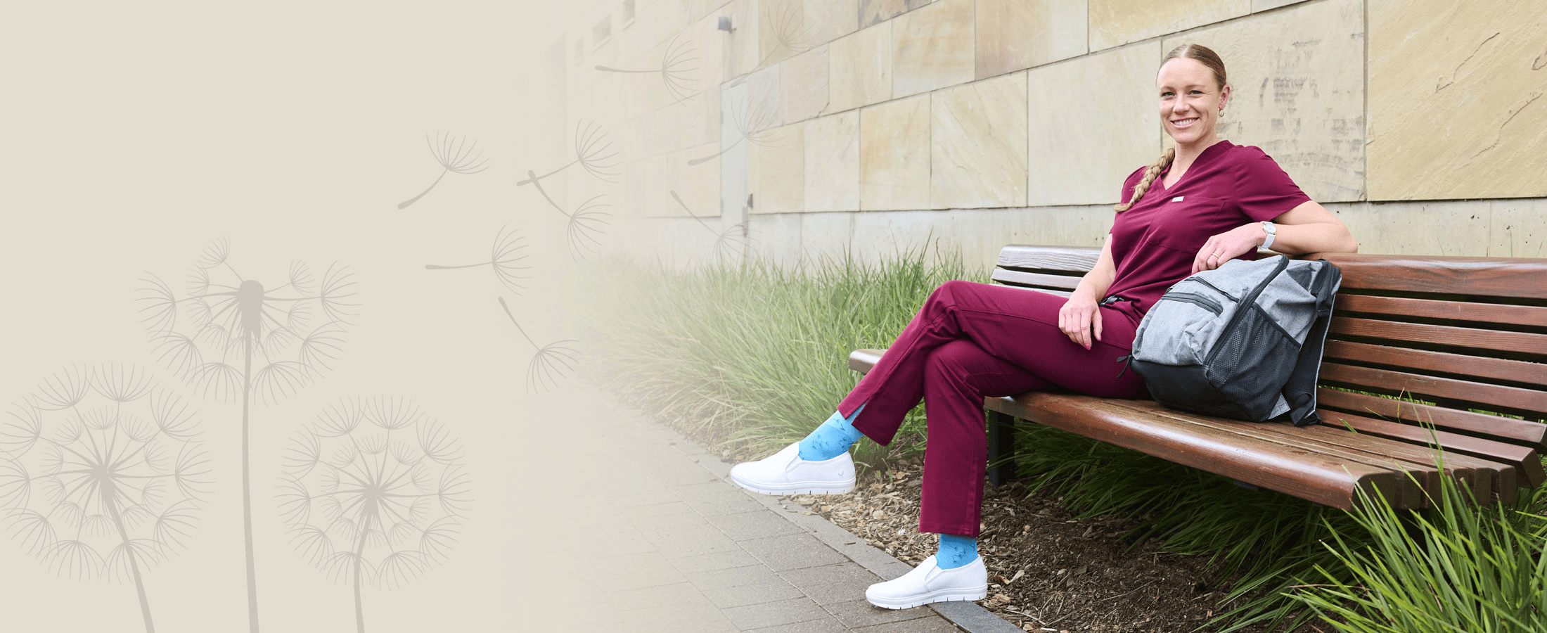 A woman sitting on a bench wearing white nursing shoes, burgundy scrubs and blue socks.