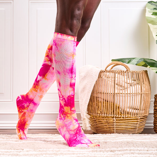 Mujer de pie con los calcetines de compresión Orange Tie Dye, de pie sobre una alfombra beige claro. Una planta y una cesta de mantas al fondo. Comprar calcetines de compresión en teñido anudado.