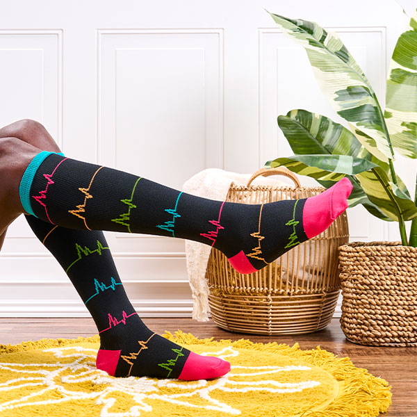 Mujer con calcetines de compresión EKG en negro, con un pie sobre una alfombra amarilla, un pie en el aire, sentada fuera del marco. Al fondo hay una planta y una cesta llena de mantas. Compra Calcetines de compresión de pantorrilla ancha en negro.