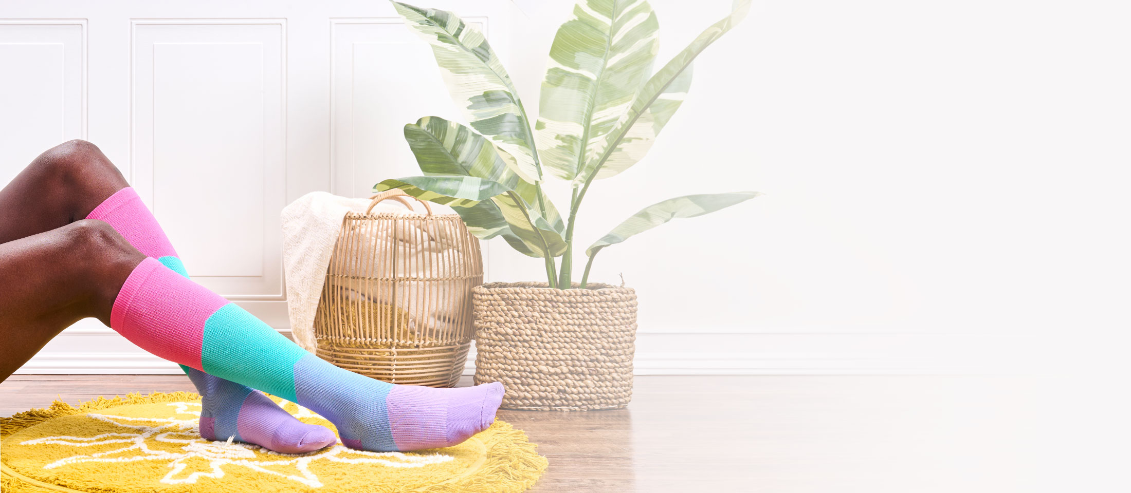 Woman wearing Color Block Bright Compression socks in multicolor, with color blocks of pink, turquoise, blue and purple, sitting out of frame with her feet on a bright yellow rug. A plant and a basket on in the background. 