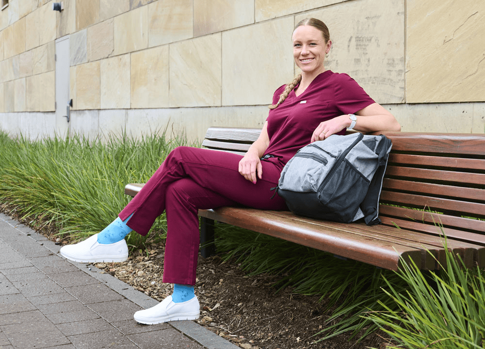 A woman sitting on a bench wearing white nursing shoes, burgundy scrubs and blue socks.